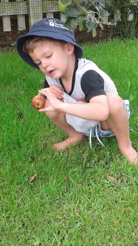 A boy in a sun hat crouching on the grass holding an apple