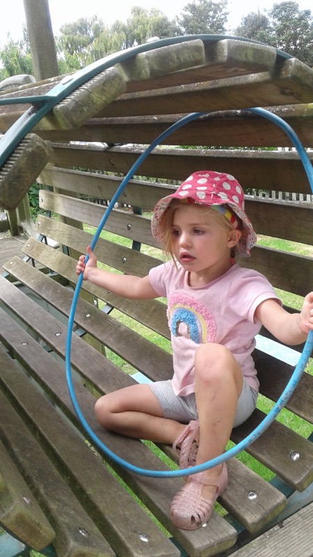 A girl in a pink sun hat sitting in a wooden climbing frame with a hula hoop
